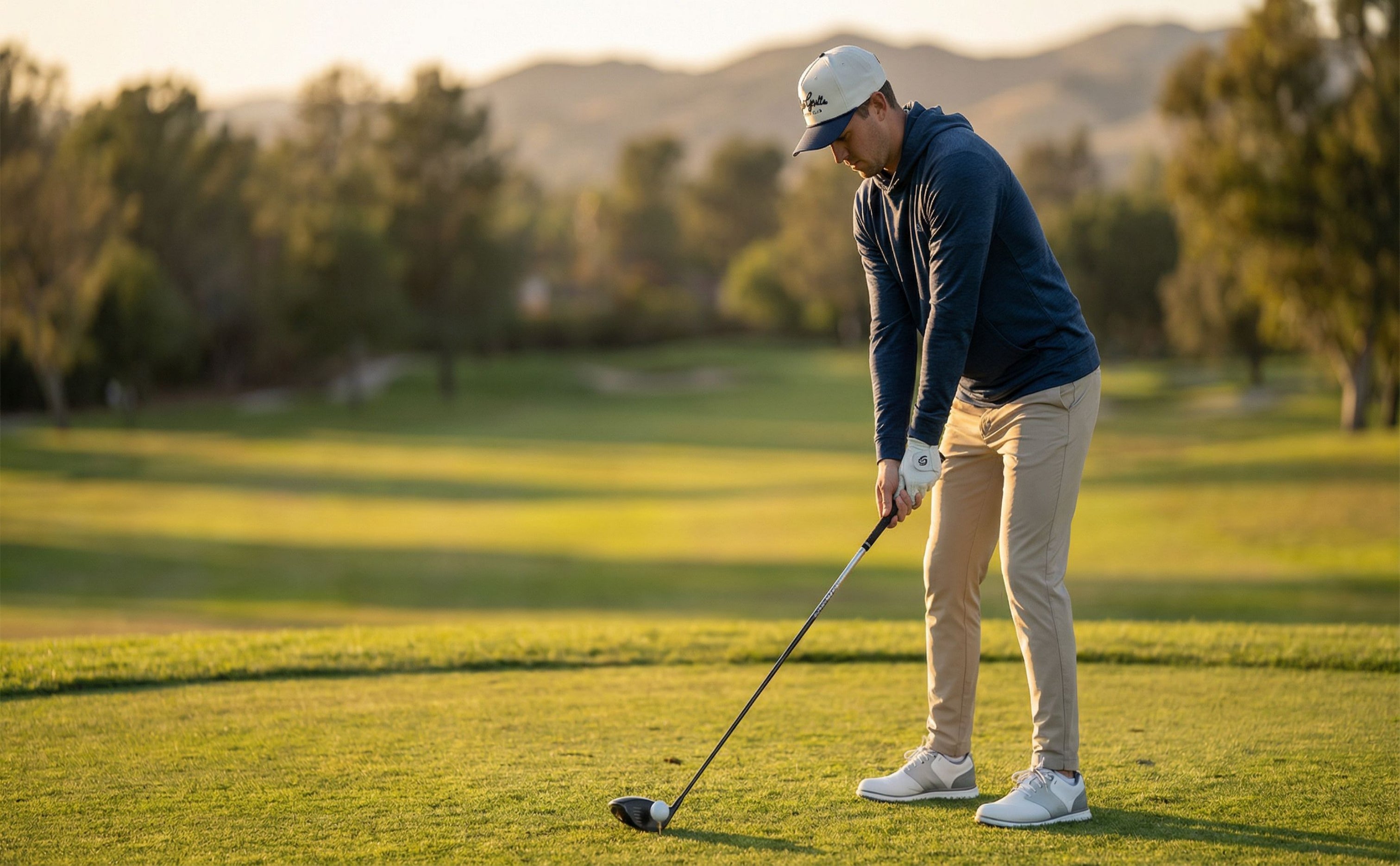 Golfer wearing a Plaid Gorilla Golf rope hat and navy hoodie addressing the ball on a tee box at golden hour